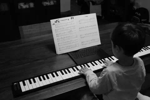 A black and white photo of a boy sat at a dining room table playing a fold-out piano. In front of him, the sheet music for a piece called 'Hocus Pocus' is propped up against a laptop screen.