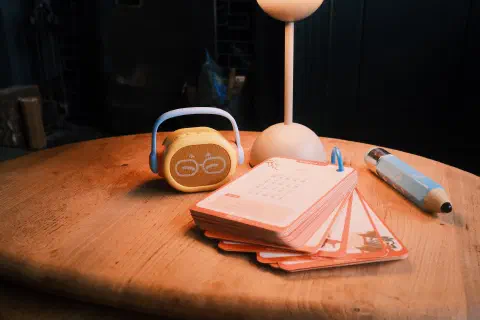 A wooden table. On the table sit a small speaker with a handle, a deck of cards on a ring binder, a large pen, and a lamp