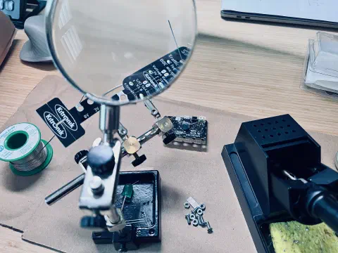 A clamp holds a magnifying glass and an electronic circuit board. Through the glass you can see the legs of a component sticking up through the board. On the desk either side of the clamp is a reel of solder and a soldering iron. A cluster of screws and nuts sit beside the base of the clamp.