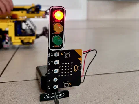 A miniature traffic light showing a red stop signal. On the stand of the taffic light, a micro:bit is mounted vertically. Wires can be seen coming from the micro:bit to a battery pack on the ground behind the traffic light. In the background a hand pushes a LEGO crane past the traffic lights.