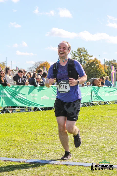 A photo of a runner crossing the finish line with a smile on their face. The runner is wearing black shoes, black shorts and a blue running top. The bib attached to their chest is sponsored by gofundme. The runners number is 4016.
