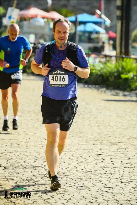 A photo of a runner in black shoes, black shorts and a blue running top. The bib attached to their chest is sponsored by gofundme. The runners number is 4016. They are runing along a cobbled road. The runner is in pain.