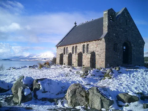A colour photo of The Church of the Good Shepherd by Lake Tekapo on South Island, New Zealand. The photo is taken in winter and the small stone church stands surrounded by snow.