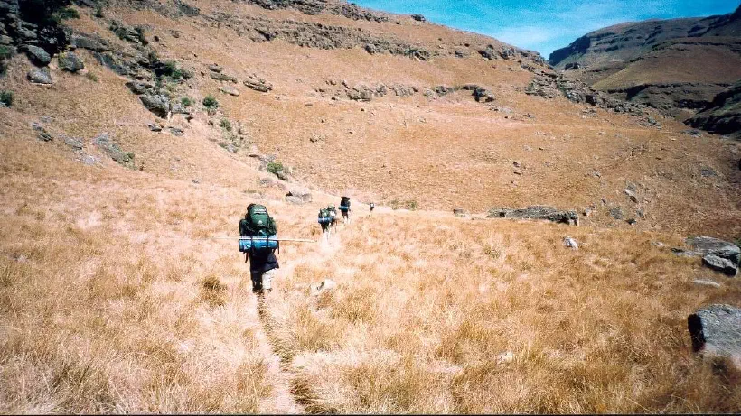A photo looking across the dry planes of the Drakensberg mountains in South Africa. A thin path leads away from the camera. Further along the path a line of hikers carry their packs up the mountain. The mountainside shows clear layers of rock in the otherwise grassy surface. The grass has been turned yellow by the dry heat of the summer sun.