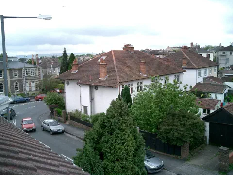 A view from the window of a house looking across the rooftops of houses opposite. A few cars are parked in the street below, but no people are visible in the photo. The sky appears cloudy but is washed out in the photograph.
