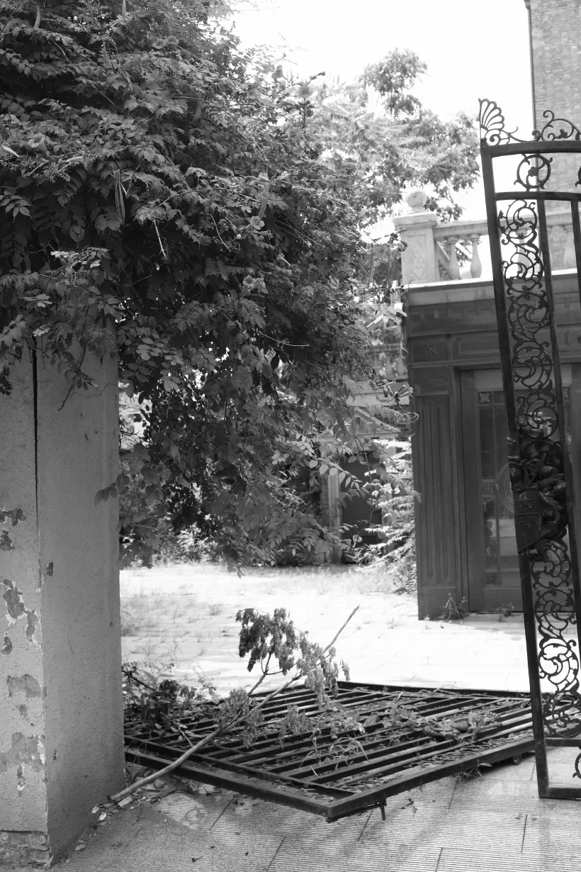 A black and white photo looking in to the front garden of a private residence in Tianjin (天津), China. The property looks run down, bushes overgrown and one of the iron gates has fallen to the ground and just left where it fell.