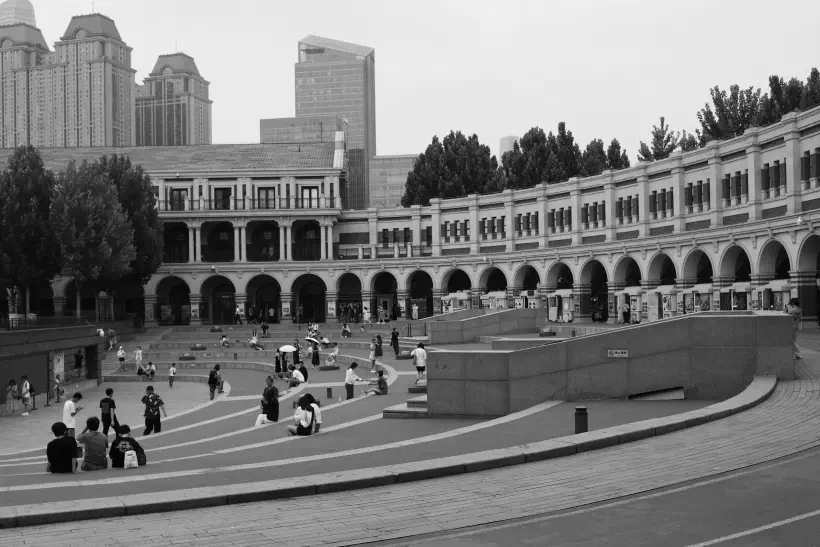 A black and white photo of Mingyuan Stadium (民园广场) in Tianjin (天津), China. The stadium has been turned into a public square and people can be seen sitting on steps that run round the inside of the stadium. Around the outer edge of what looks like a running track, an arched covering provides shade from the heat. The stadium feels like it was once a hive of activity, today it feels as if there is something missing.
