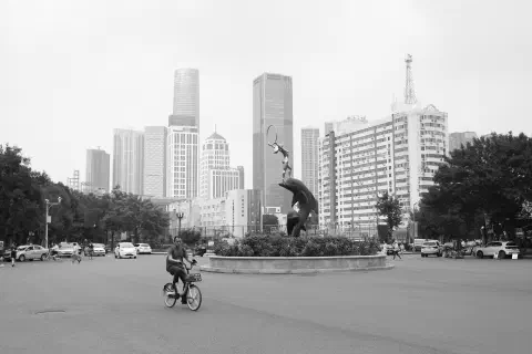 A black and white photo of a roundabout in Tianjin (天津), China. In the foreground, a lone cyclist occupies the roundabout on a rental bike. A statue stands in the middle of the roundabout.Tall skyscrapers line the horizon in the distance.