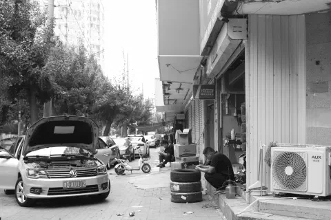 A black and white photo of a street in Tianjin (天津), China. On the left of the image a car sits parked, the passenger side door open and the bonnet (hood) propped up. On the right, a mechanic sits by a pile of three tyres on the steps leading to his workshop. His focus is on the phone in his hands. In the distance, a man sits on the steps of the next shop. He is also looking at his phone.