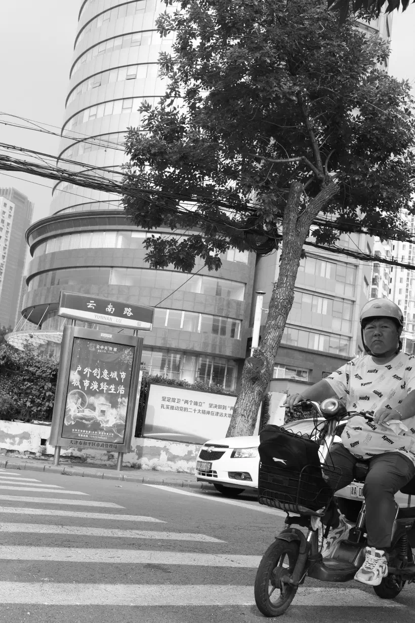 A black and white photo taken on the streets of Tianjin (天津), China. The photo is take from low down, looking up across a pedestrian crossing.  A man on an electric scooter looks purposfully at the camera as he enters the crossing. On the far side of the street, a curved glass building rises up to the top of the frame.