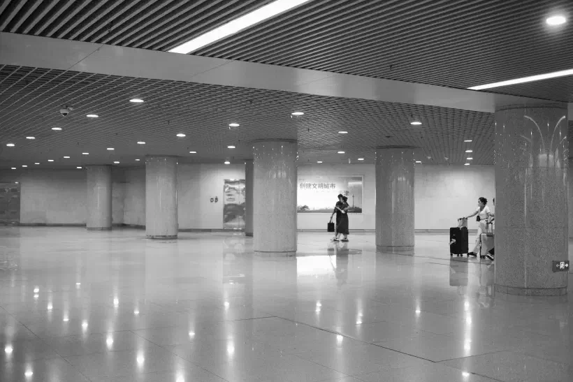 A black and white photo of the subway station in Tianjin (天津), China. The station is empty save for three people making their way across the hall with their lugguage. The bright ceiling lights reflect in the polished floor.