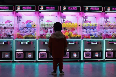 A young child stands with their back to the camera, looking at a row of arcade machines. The turquoise and pink lighing from the machines reflects in the pollished floor.