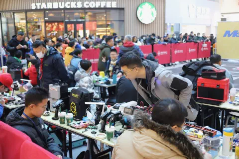 A crowded area in a shopping mall in China. A row of tressle tables is covered with tools, electronics, cars and sugary drinks. At the tables, people sit repairing their cars. In the background a crowd stands up against a barrier in front of a Starbucks. They apear to be waiting to watch an event.