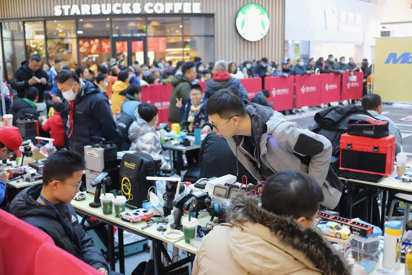 A crowded area in a shopping mall in China. A row of tressle tables is covered with tools, electronics, cars and sugary drinks. At the tables, people sit repairing their cars. In the background a crowd stands up against a barrier in front of a Starbucks. They apear to be waiting to watch an event.
