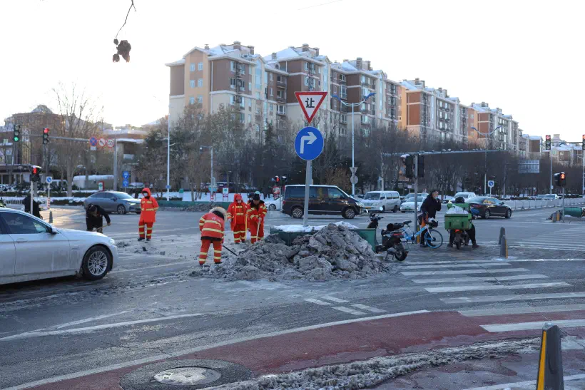A photo of a road junction in China nearest to the camera is a right hand turn lane. Just the other side of the lane is a pile of snow as workmen take advantage of the red traffic lights to clean snow from the main carriageway.