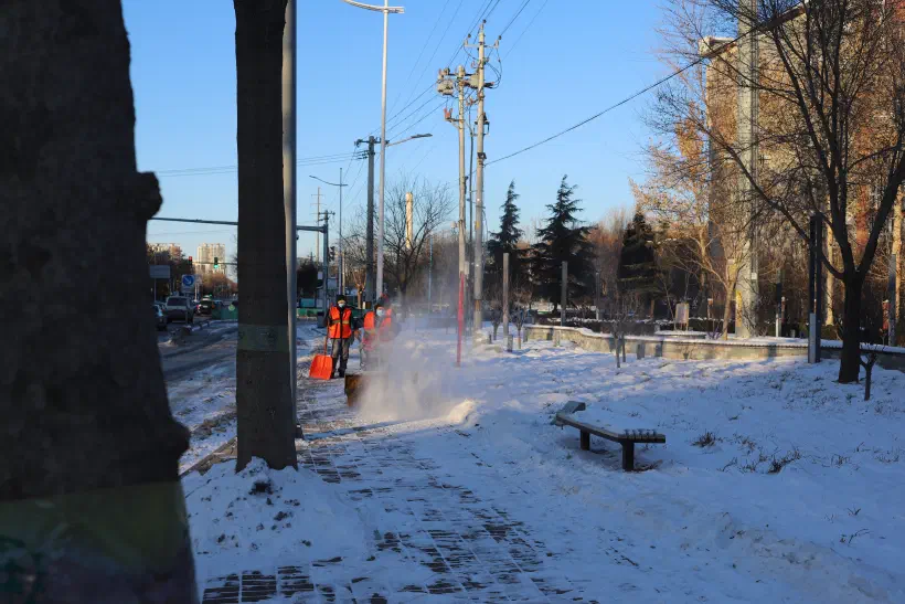 Looking down a snow covered footpath at the side of a road. In front, a hand-pushed snow plow churns snow and ice off the sidewalk.
