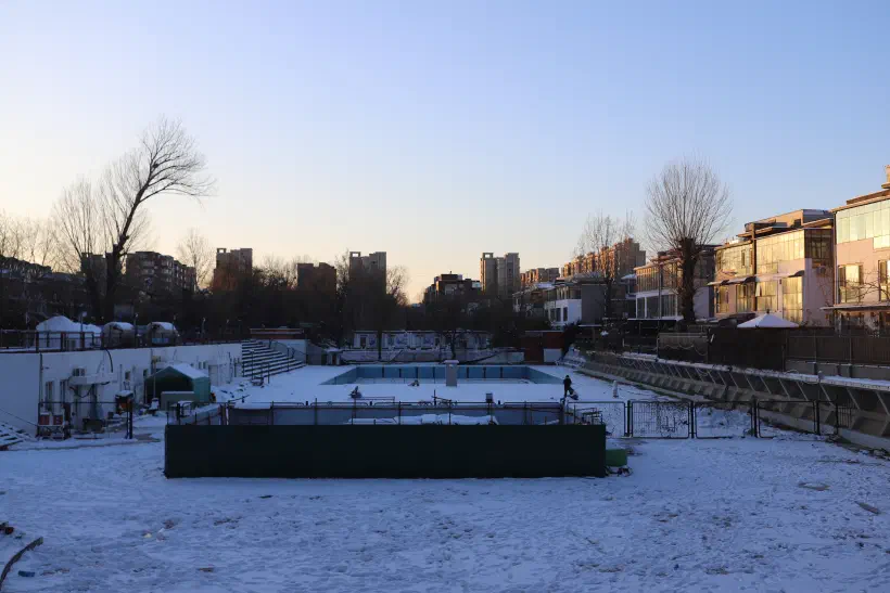 A photo looking over a disused swimming pool. Surrounding the pool are residential buildings the pool and surrounding walkways are covered in snow.