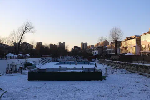 A photo looking over a disused swimming pool. Surrounding the pool are residential buildings the pool and surrounding walkways are covered in snow.