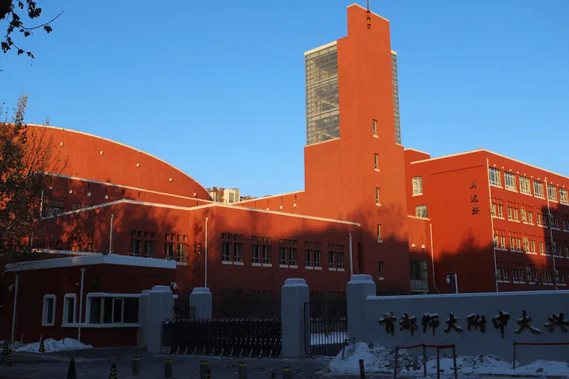 A photo of a red building brightly lit in the morning sunshine. The sign on the gate to the school indicates that this is DaXing Middle School in Beijing.