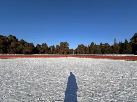 A wide angle photo looking across flat, snow-covered ground. A red wall extends from the sides of the image to a vanishing point in the centre of the phtoo. Trees poke above the top of the wall, standing in contrast to the bright blue sky. The shadow of the the photographer is visible in the front of the image.
