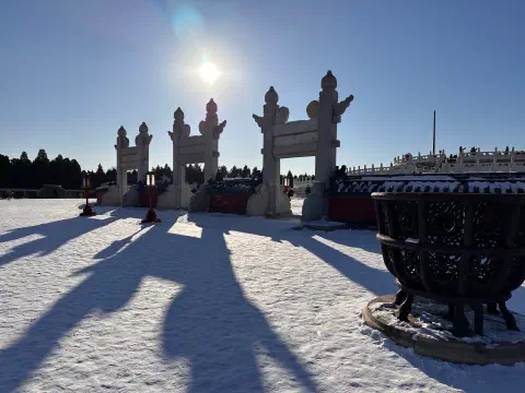 A photo of a wintery scene inside the Temple of Heaven in Beijing. The sun is low in a cloudless sky. There stone gateways stand at the entrance to the circular mound, their long shadows extending all the way to the photographer.