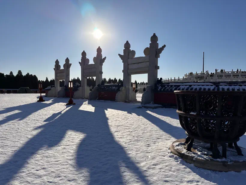 A photo of a wintery scene inside the Temple of Heaven in Beijing. The sun is low in a cloudless sky. There stone gateways stand at the entrance to the circular mound, their long shadows extending all the way to the photographer.