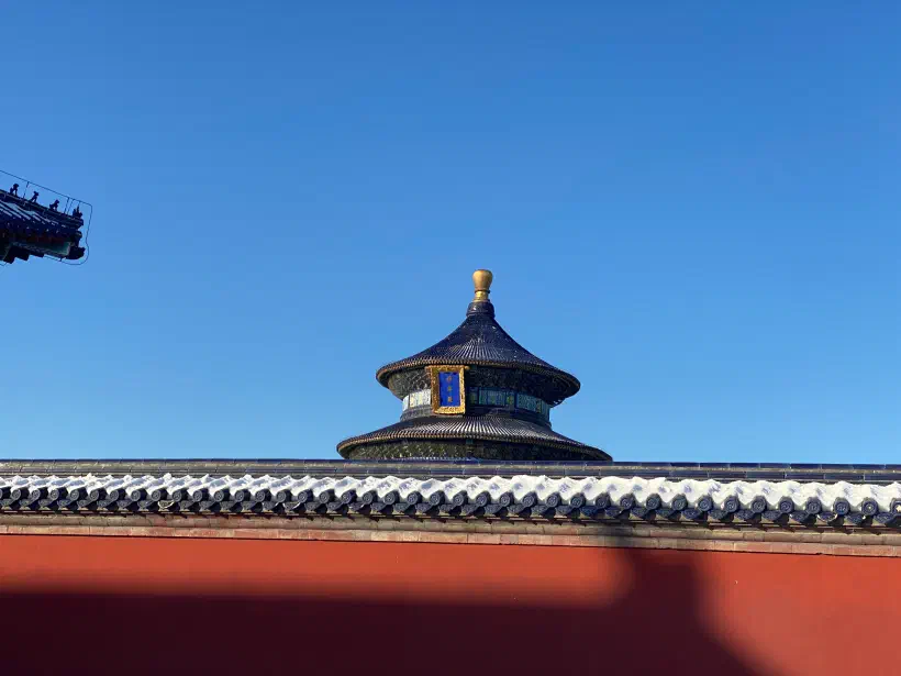 A photo looking up over a red wall in the Templ of Heaven, Beijing. Snow covers the top of the wall. Behind it, the circular roof-top of a temple. The sky is blue with no clouds.