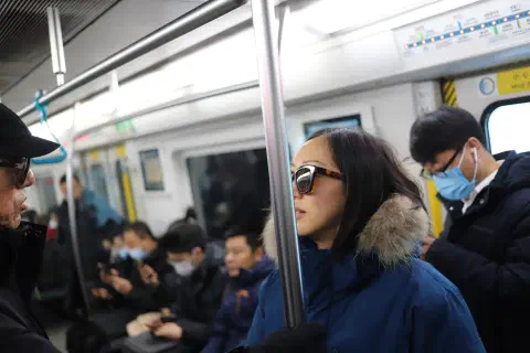 A photo inside a Beijing subway car. Most passengers are wearing face masks. A person stnds looking to the left of the image wearing sunglasses.