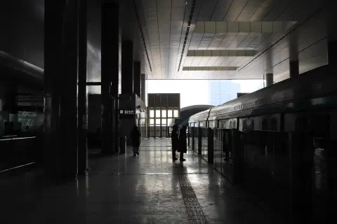 A photo looking along a covered subway platform in Beijing China. The bright sunlight from the opening at the far end of the platform silhouettes two passengers standing on the platform.