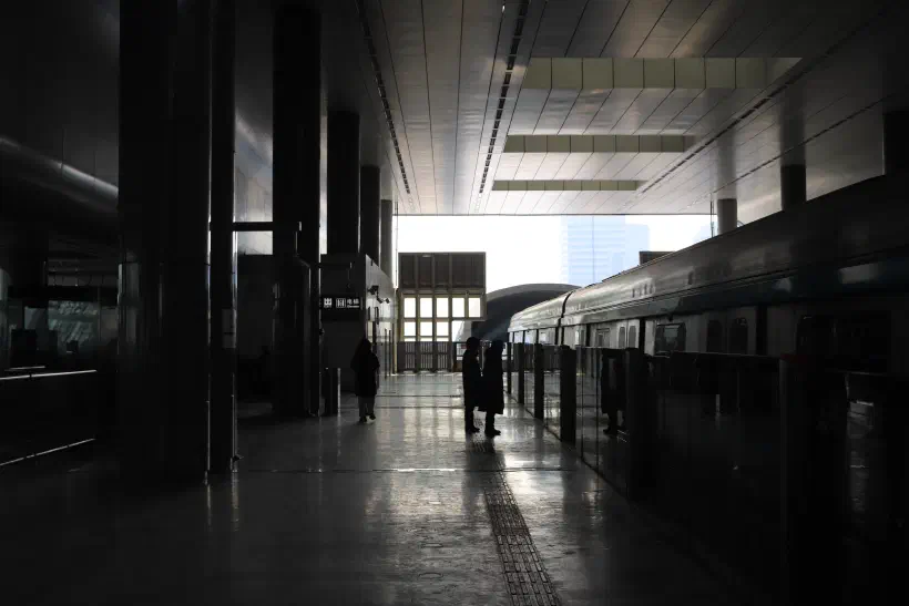 A photo looking along a covered subway platform in Beijing China. The bright sunlight from the opening at the far end of the platform silhouettes two passengers standing on the platform.