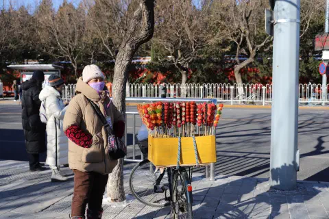A street vendor selling candied fruit from a small cart in Beijing. The vendor is wrapped up warm, hands in pockets to protect them from the cold. The street behind them is empty of vehicles.