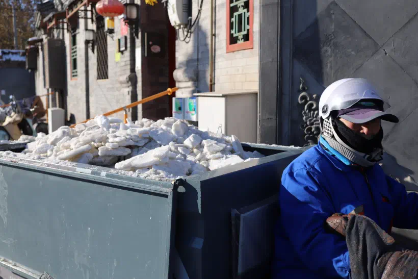 A small motorised tricycle with an open bed removes sheets of snow and ice in Beijing. The driver is wrapped up warm in a blue overall. Their face isn't visible, wrapped up and protected from the cold.