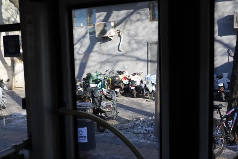 A view through the window of a moving bus looking onto a Beijing street. Buildings are grey, alongside them a row of mopeds and bicycles.