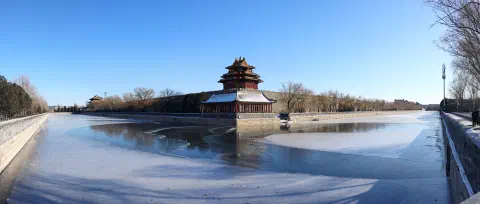 A panorama looking through trees onto the north-west corner of the Forbidden City in Beijing. The frozen moat is just visible before the city walls. The ground before the moat is completely covered in snow.
