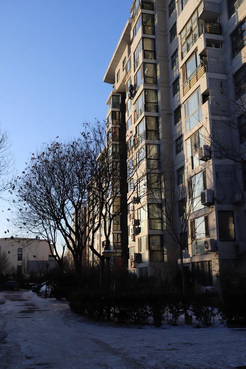 A photo of a high-rise apartment building in Beijing. The building is lit with the warm evening sun. On the road in front of the building, a covering of snow.