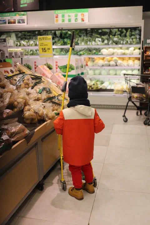 A child pushes a small supermarket trolley around the isles of a supermarket. To their left an array of root vegetables. In front of them, fresh green leaves.