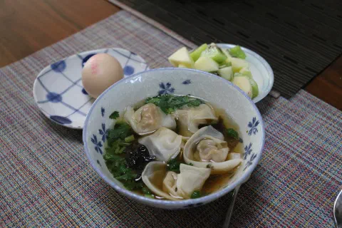 A bowl of wonton and broth alongside a boiled egg and a plate of fruit.