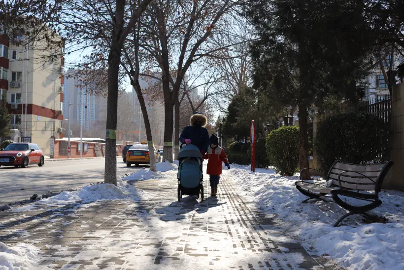 A family walk along the snow covered sidewalk along a Beijing street.