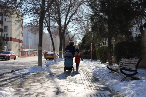 A family walk along the snow covered sidewalk along a Beijing street.