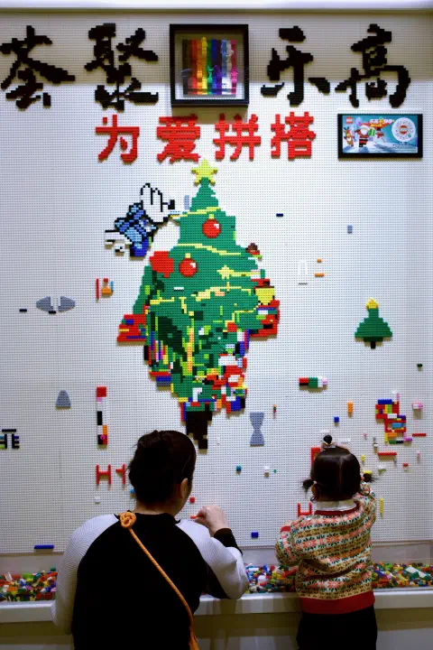Two people place LEGO bricks on a wall of LEGO base plates. The wall is decorated with Chinese characters, a Christmas tree and small little creations out of colourful LEGO bricks.