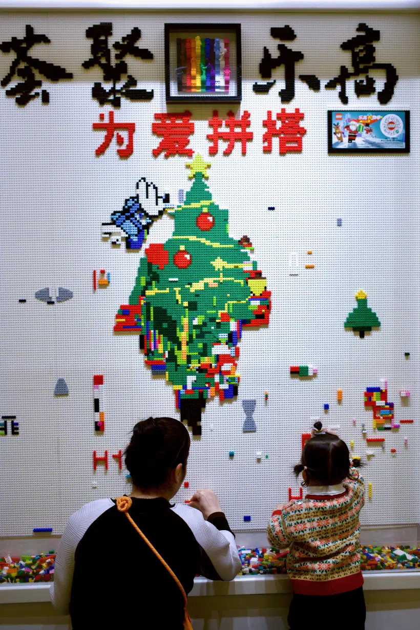 Two people place LEGO bricks on a wall of LEGO base plates. The wall is decorated with Chinese characters, a Christmas tree and small little creations out of colourful LEGO bricks.