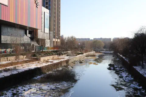 A river flows past a shopping mall in Beijing, China. The banks of the river are covered in snow and ice.