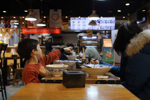 Two people sit at a restaurant table. In front of them two large bowls. They are looking away from the camera towards the restaurant counter.