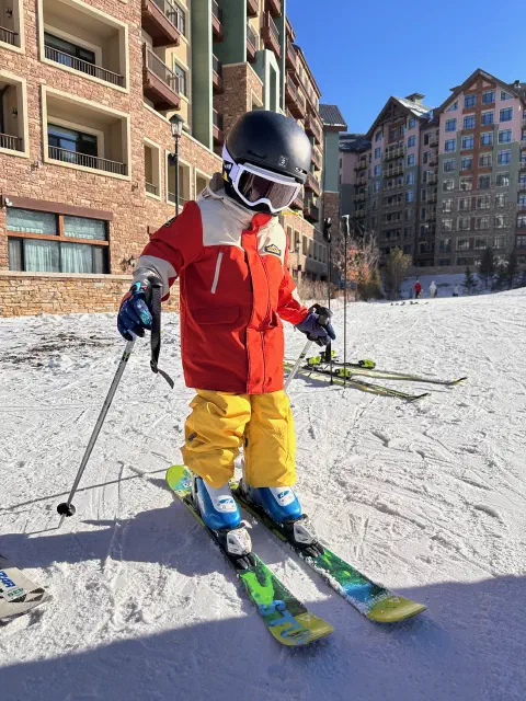 A young child pushes off on skiis. Behind them ski challets line the slopes. The child is wearing an orange jacket and yellow trousers.