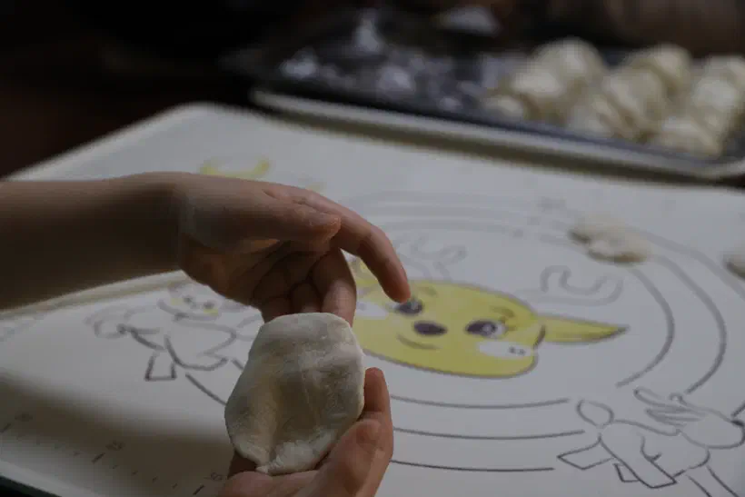 Young hands hold up a freshly rolled dumpling skin. The skin is not round but somewhat oval in shape.