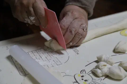 A photo of a hands using a plastic knife to cut pieces of fresh dough for Chinese dumplings.