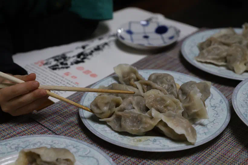 A person (out of frame) uses chopsticks to pick up a dumpling from a plate of boiled dumplings in the middle of a table.