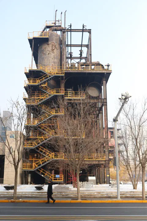 An industrial-looking building stands against a grey sky. In front of the building, a person walks along the road. The building itself is rusty, with a yellow, freshly painted staircase down the outside. The trees are all bare, and it looks like winter.