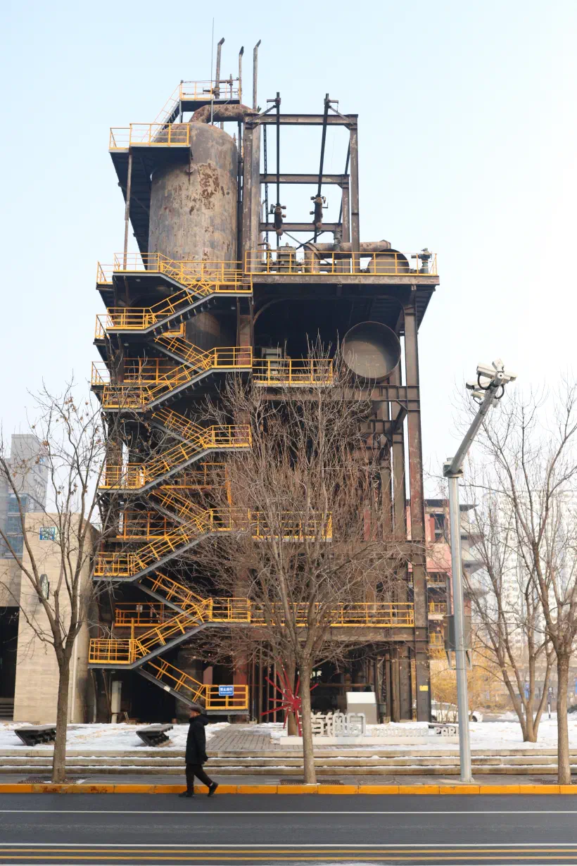 An industrial-looking building stands against a grey sky. In front of the building, a person walks along the road. The building itself is rusty, with a yellow, freshly painted staircase down the outside. The trees are all bare, and it looks like winter.