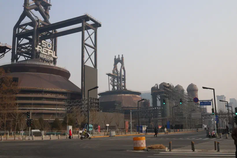 on the side of an old rusty factory building a yellow staircase winds its way up to the top. On the road, a lone person walks past the building.